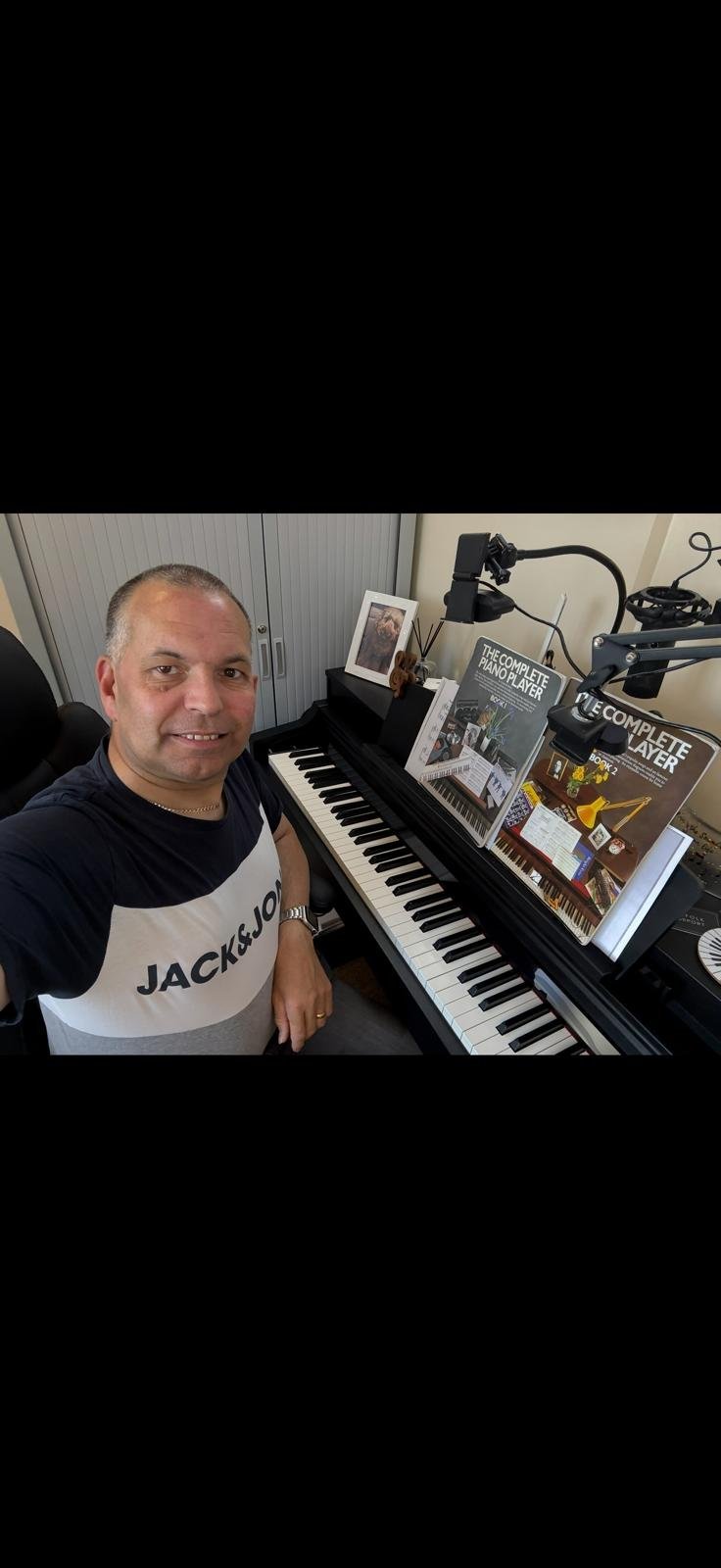 Graham Woods, music teacher, smiling at his home studio digital piano with music books and microphone set up for a lesson